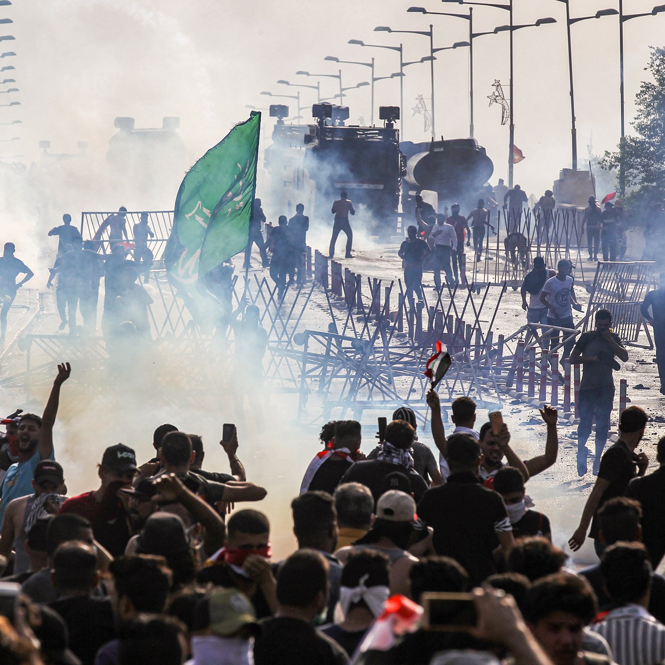 L'image montre une scène de tumultes ou de manifestations. On y voit un grand groupe de personnes rassemblées, brandissant des drapeaux et levant les bras. Il y a des barrages en bois et de la fumée, probablement causée par des gaz lacrymogènes. À l'arrière-plan, des véhicules blindés sont visibles, renforçant l'idée d'une situation tendue. L'atmosphère est chargée et chaotique, avec des manifestants qui semblent déterminés et en mouvement.