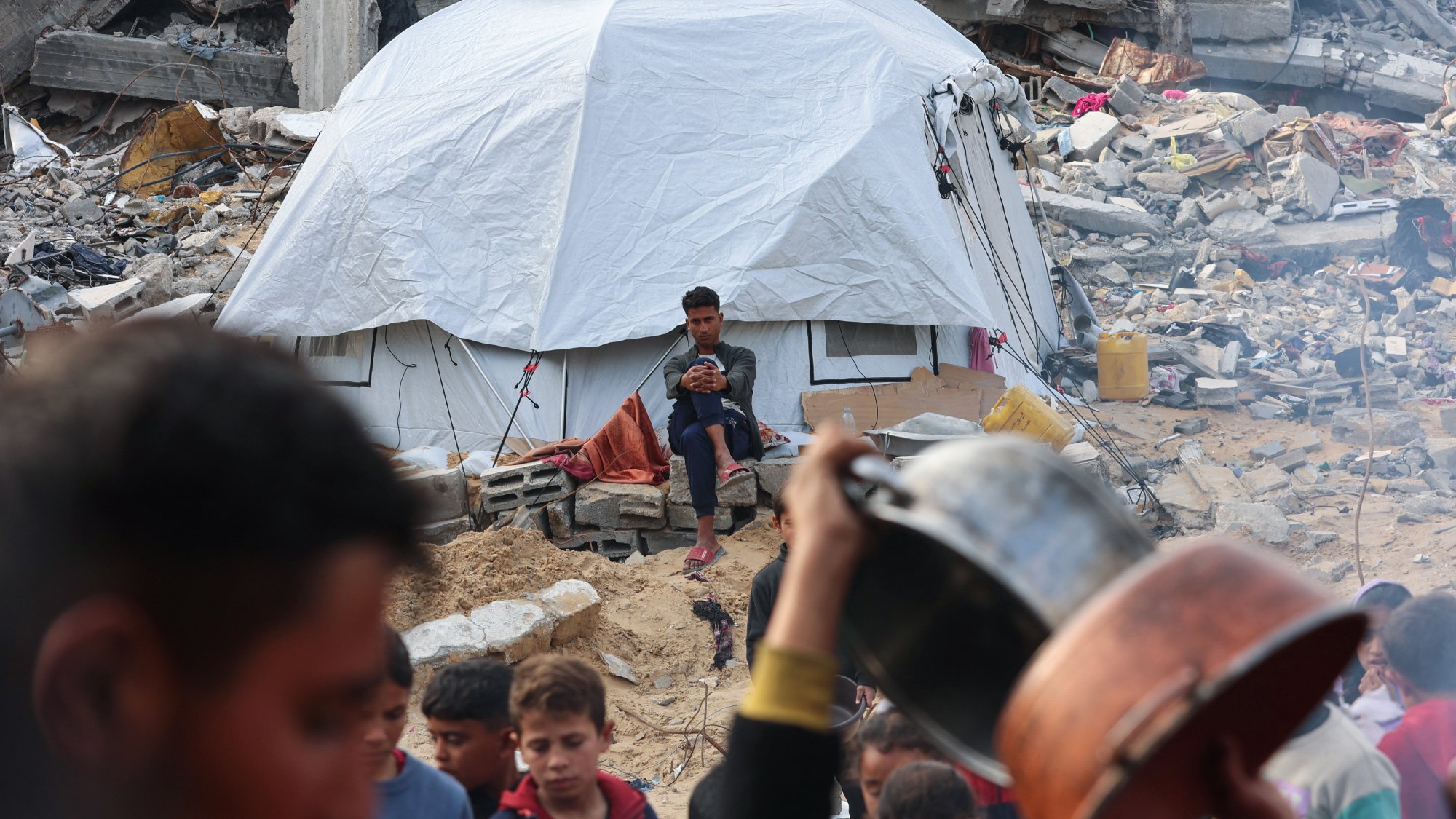Nell'immagine si vede una tenda bianca in un contesto di distruzione, con rovine e macerie intorno. Un ragazzo è seduto davanti alla tenda, mentre in primo piano ci sono persone che si muovono e tengono stoviglie. L'atmosfera sembra di emergenza e difficoltà.