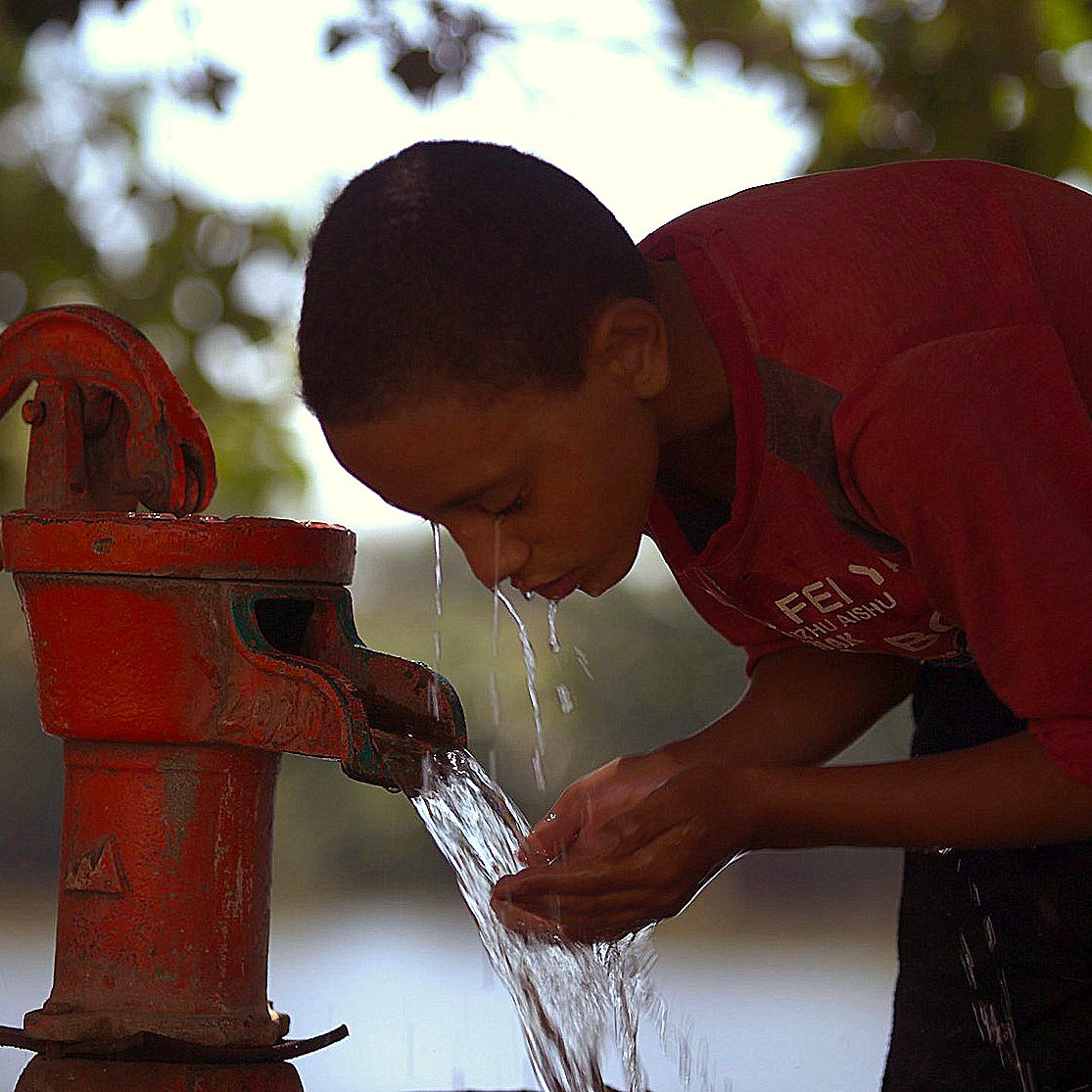 في الصورة، يظهر صبي صغير bent forward ليشرب من نافورة مياه. يستخدم يديه لالتقاط المياه المتدفقة من الحنفية الحمراء. الخلفية تضم شجرة وأجواء طبيعية هادئة، مما يعكس مشهدًا من الحياة اليومية حيث يسعى الطفل للحصول على الماء. الإضاءة في الصورة تضفي شعورًا بالسلام والحياة البسيطة.
