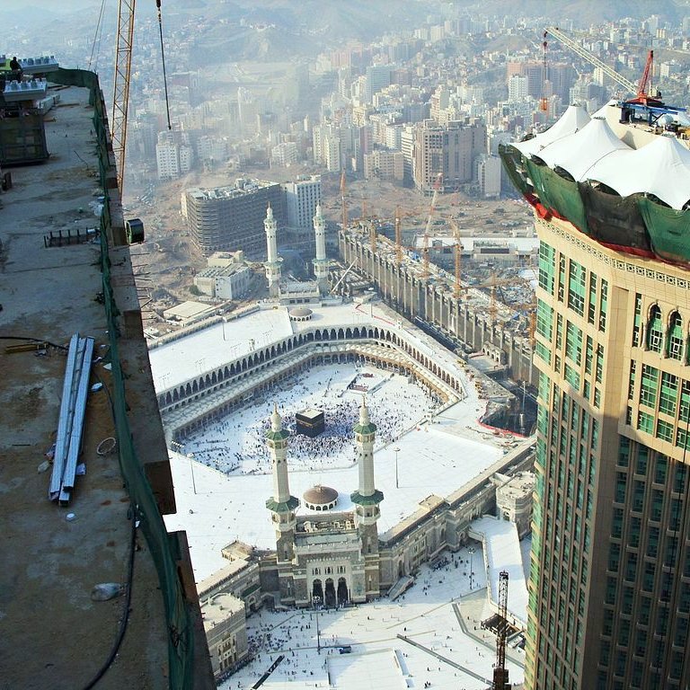 L'image montre une vue aérienne de la Kaaba à La Mecque, en Arabie Saoudite. On peut voir le complexe du Haram qui comprend la mosquée entourant la Kaaba. Il y a de nombreux pèlerins qui s'y trouvent, et l'environnement urbain avec des bâtiments modernes en arrière-plan. On aperçoit également des travaux de construction en cours sur le côté, suggérant une expansion des infrastructures autour du site sacré. Le ciel est légèrement brumeux, ce qui donne une ambiance particulière à la scène.