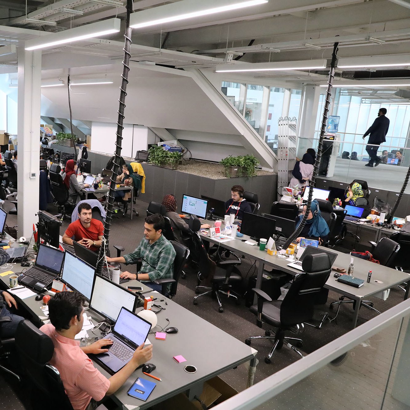 الصورة تظهر مساحة عمل مكتبية حديثة. تتميز بتصميم مفتوح حيث يجلس العديد من الأشخاص أمام شاشات الكمبيوتر. توجد طاولات بتوزيع غير منتظم، وبعض الأشخاص يعملون بشكل فردي بينما الآخرون يتعاونون معًا. يبدو الجو نشطًا وحيويًا، مع وجود نباتات في الأرجاء تضفي لمسة من الطبيعة. هناك أيضًا لافتات وأدوات مكتبية متنوعة، مما يدل على بيئة عمل مهنية حديثة.