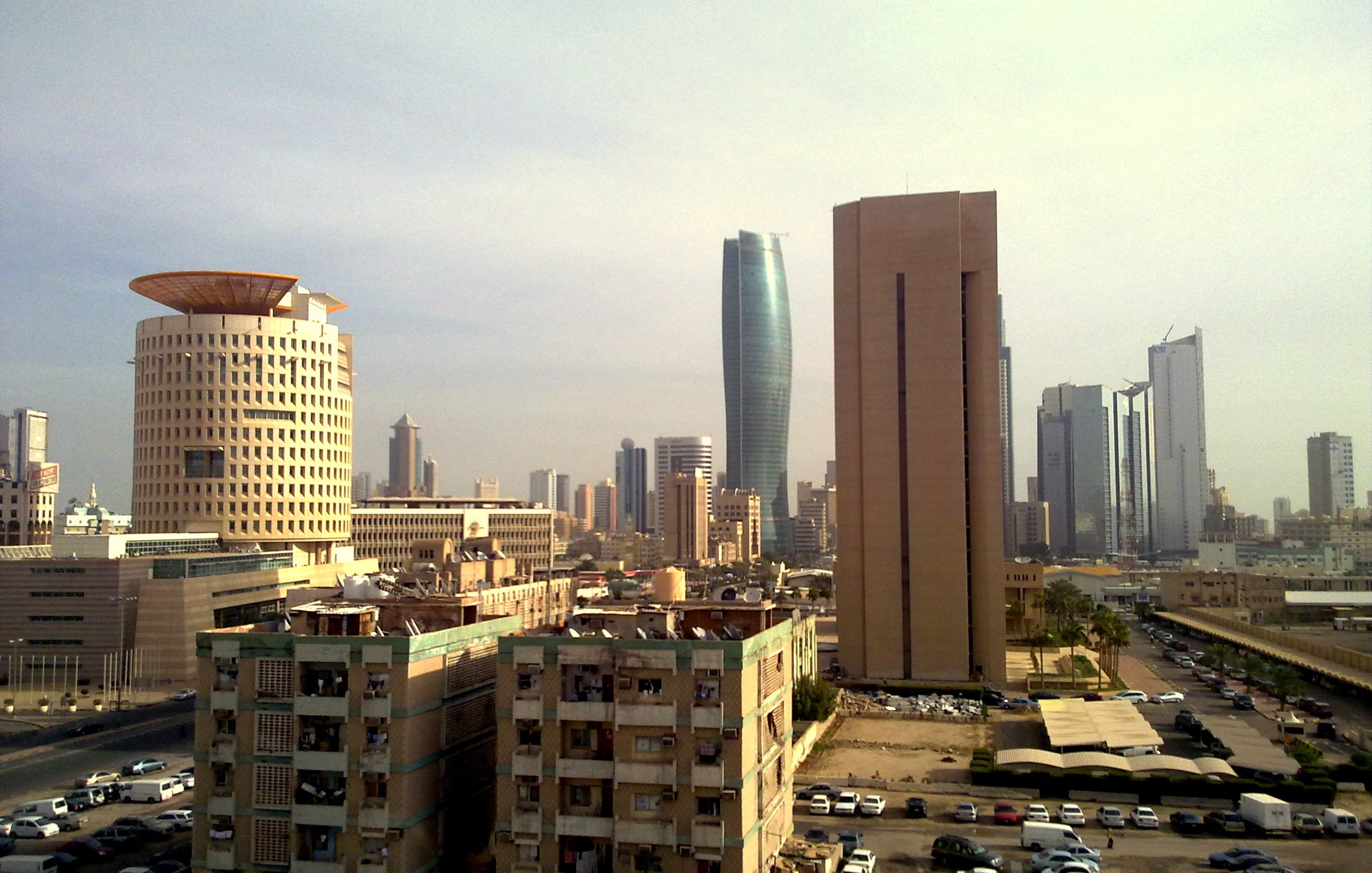 L'image montre une vue urbaine d'une grande ville moderne. On peut apercevoir plusieurs gratte-ciel aux architectures variées, avec des bâtiments à la fois anciens et contemporains. À gauche, un bâtiment au design unique, semblable à un tambour, et à droite, une structure plus élancée et moderne. En bas, on voit des immeubles vieillissants et des espaces de stationnement. Le ciel est légèrement nuageux, donnant une atmosphère calme à la scène. L'ensemble dépeint un mélange d'éléments historiques et modernes d'une métropole dynamique.