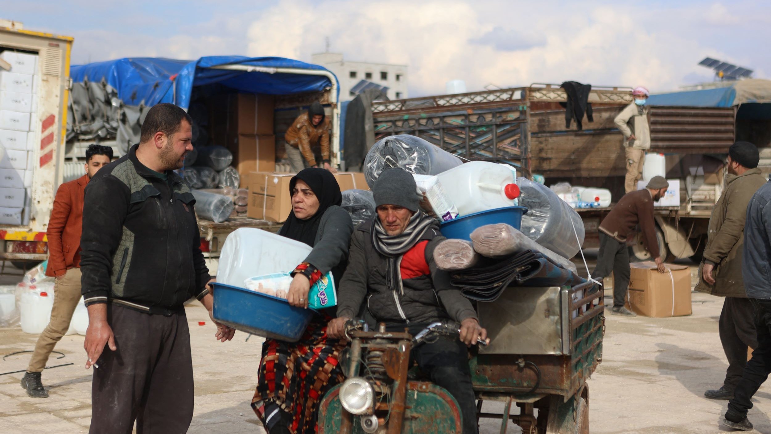 Nell'immagine si vede una scena di un'area di stoccaggio o mercato. Ci sono alcune persone che sembrano occupate a scaricare e trasportare materiali. Un uomo e una donna sono in primo piano: l'uomo indossa abiti scuri e tiene una cassetta, mentre la donna è vestita con un lungo mantello scuro e apparente una certa preoccupazione sul volto. Dietro di loro, ci sono merci come bottiglie di plastica e tappeti, oltre a veicoli e altri uomini che svolgono attività di carico e scarico. L'atmosfera sembra essere quella di un'operazione di supporto umanitario.