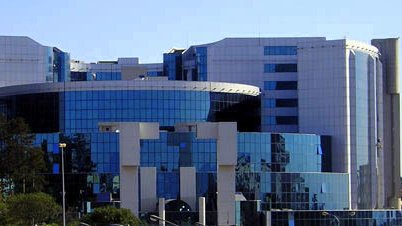 L'image montre un bâtiment moderne au design architectural contemporain. La façade est principalement en verre, ce qui reflète la lumière et les bâtiments environnants. Des formes géométriques et des courbes sont bien mises en valeur, et on peut distinguer des éléments en métal. Le paysage autour est agrémenté d'arbres et de plantes, suggérant que le bâtiment est situé dans une zone urbaine. L'ensemble dégage une impression de sophistication et de modernité.