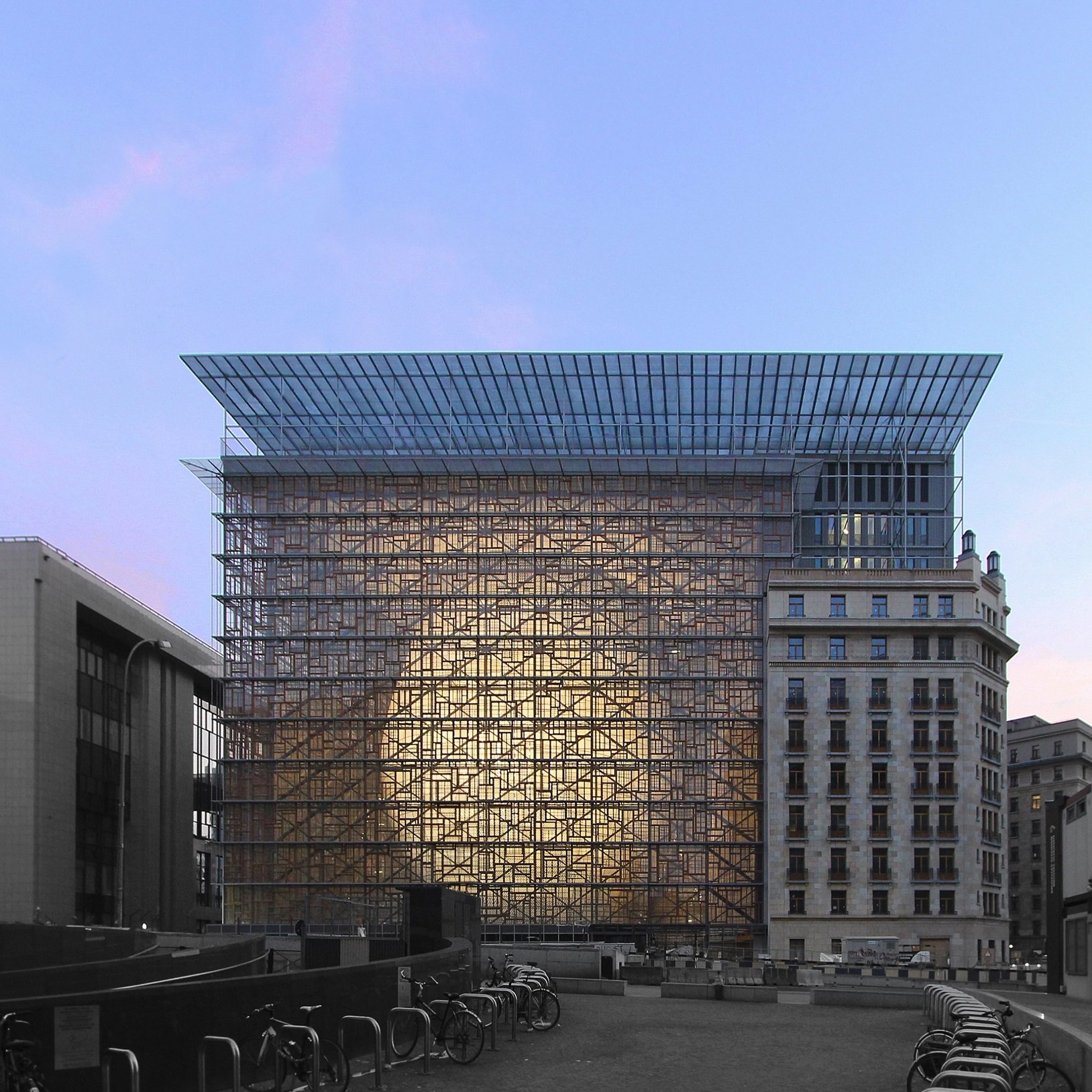 L'image montre un bâtiment moderne avec une structure en verre et en métal, se dressant au centre d'un environnement urbain. La façade réfléchissante crée un effet de transparence et de lumière, tandis que le ciel présente des nuances de bleu et de rose, suggérant un moment au crépuscule. Au premier plan, on aperçoit des vélos alignés, ajoutant une touche de vie à la scène. À gauche, il y a un autre bâtiment avec un style architectural plus traditionnel, contrastant avec la modernité du bâtiment principal.