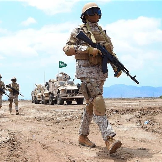 الصورة تظهر مجموعة من الجنود في زي عسكري، يسيرون على الأرض في منطقة صحراوية أو شبه صحراوية. الجنود يرتدون خوذات واقية ودرع، ويبدو أنهم في حالة استعداد. في الخلفية، توجد مركبات عسكرية مدرعة، ويظهر العلم السعودي. السماء تبدو صافية مما يشير إلى يوم مشمس. الجو العام يوحي بالتدريب العسكري أو المناورات.