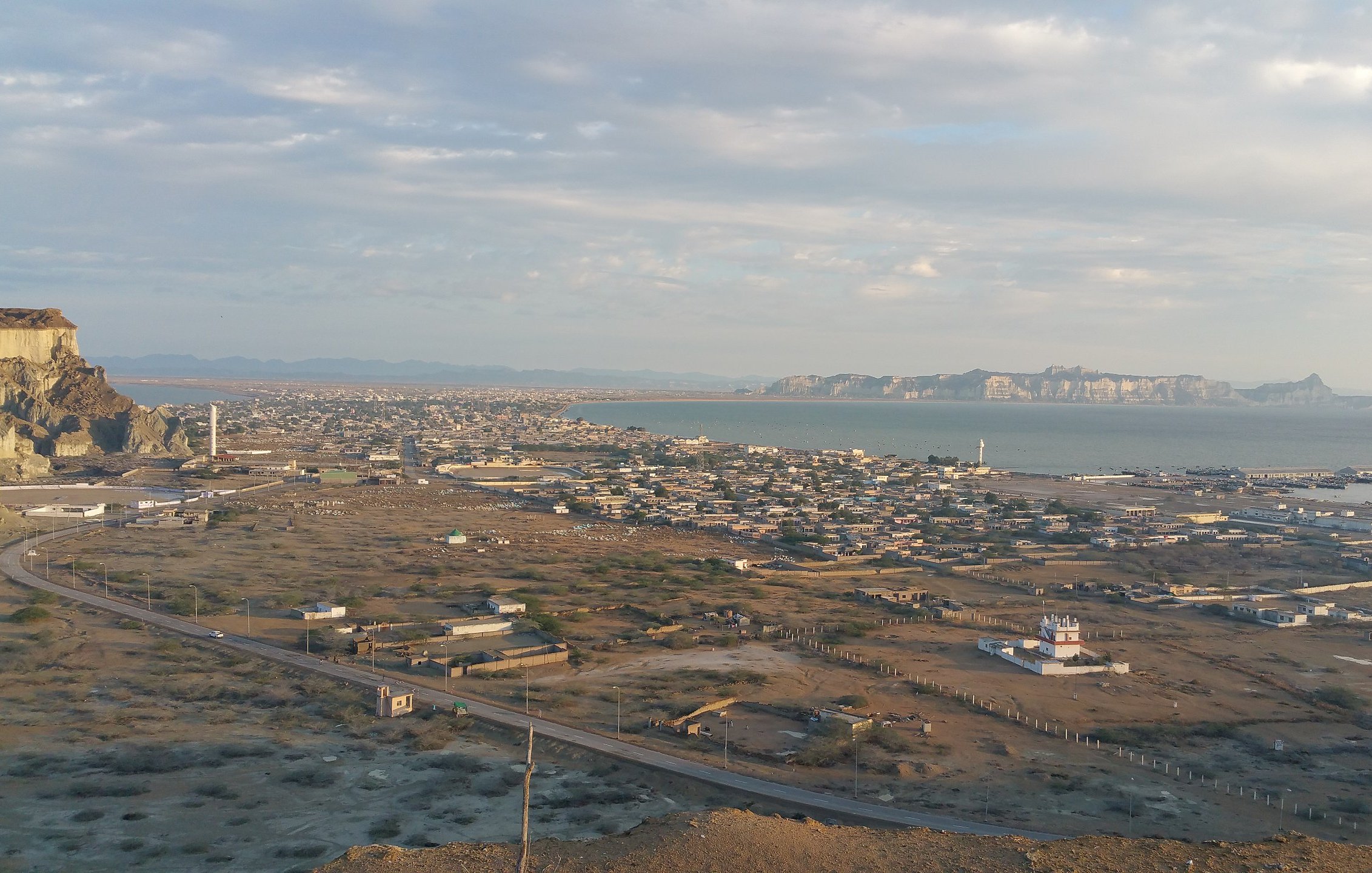 L'image montre un paysage côtier avec une vue panoramique. On peut voir une ville située au bord de la mer, entourée de collines et de montagnes à l'horizon. La mer est calme, et des structures côtières, comme des docks ou des bateaux, sont visibles. Le ciel est partiellement nuageux, ce qui donne une belle lumière à la scène. On observe également des zones désertiques ou semi-désertiques, avec des routes qui serpentent à travers le paysage.