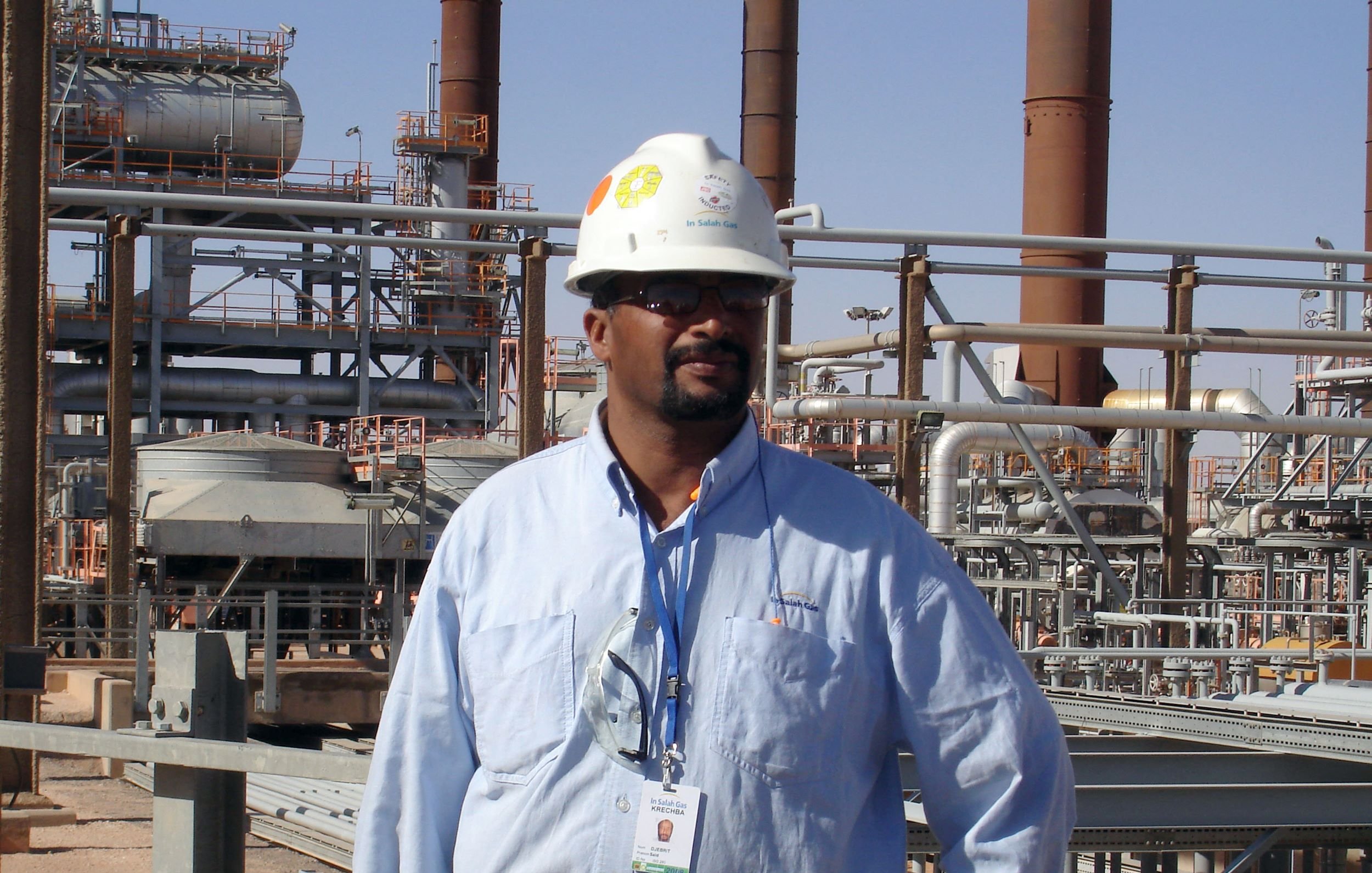 L'immagine mostra un uomo che indossa un casco di sicurezza bianco e una camicia azzurra. Si trova in un ambiente industriale, con impianti e tubazioni sullo sfondo. L'atmosfera sembra quella di un sito di produzione o di una raffineria, con diverse strutture metalliche e torri visibili. L'uomo appare fiducioso e professionale mentre si pone di fronte all'obiettivo.