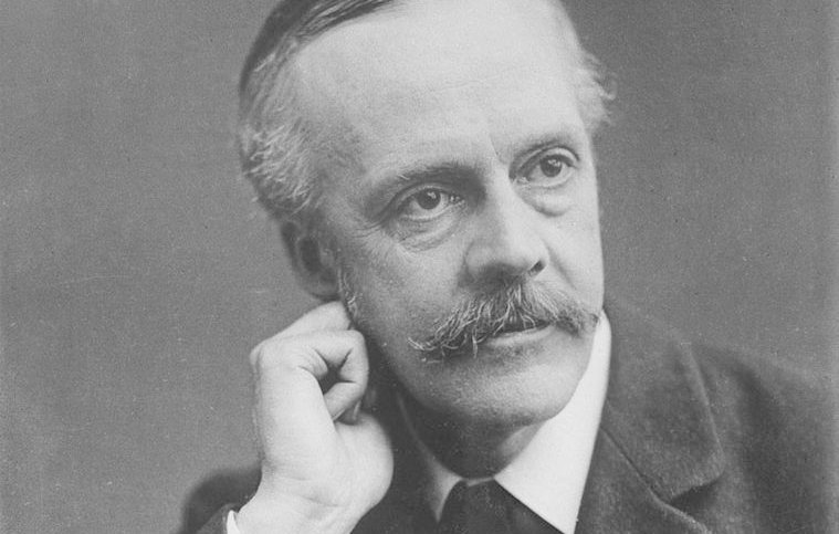 L'image montre un homme en costume vêtu d'une veste sombre et d'une cravate. Il a des moustaches bien entretenues et les cheveux légèrement grisonnants. Il pose en penchant légèrement la tête sur la main, affichant une expression pensive. Le fond est neutre, mettant en valeur son visage et son regard pensif.