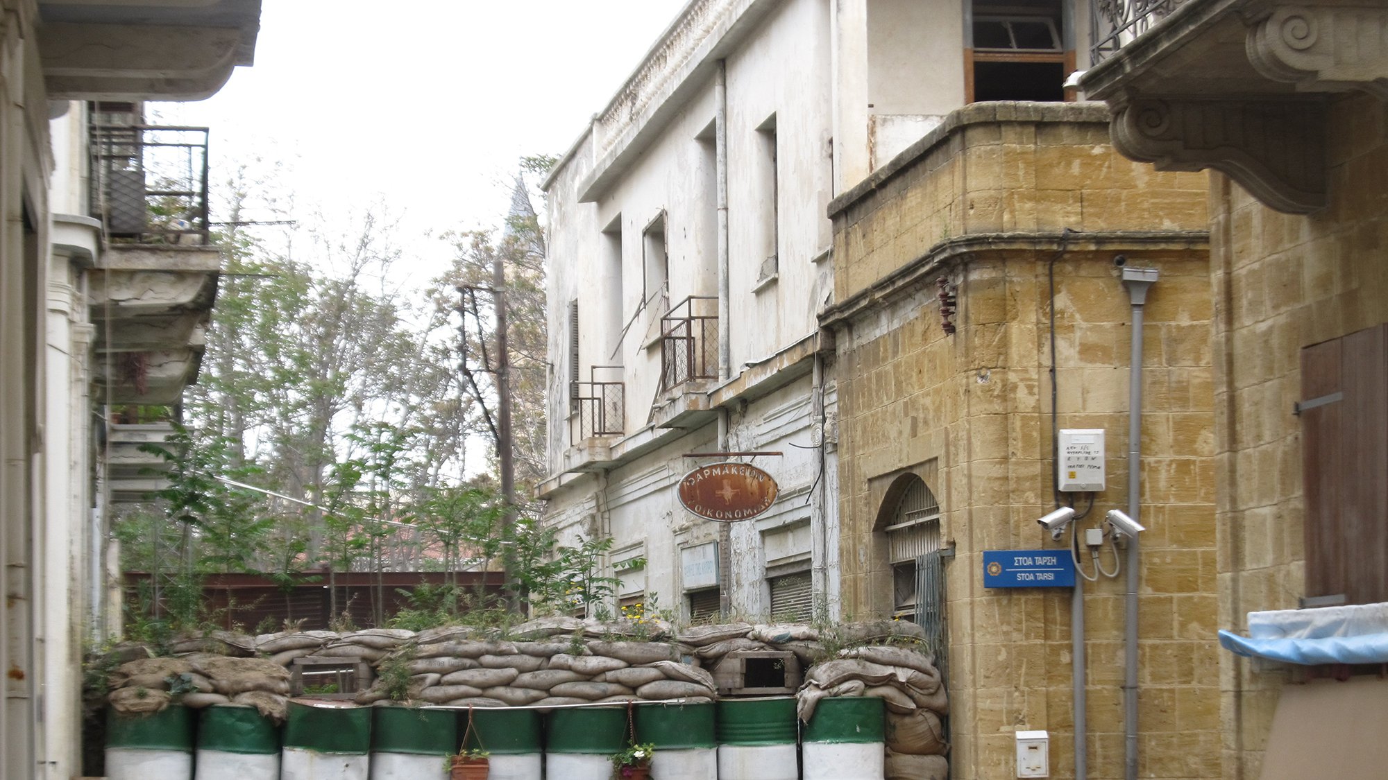 الصورة تظهر شارعاً ضيقاً في مدينة قديمة، حيث توجد مباني قديمة ذات تصميم معماري تقليدي. في نهاية الشارع، هناك حاجز مصنوع من أكياس الرمل وبراميل، مما يشير إلى وجود خطط للحماية أو الأمن. بعض النباتات تنمو حول الحاجز، مما يضفي لمسة من الحياة على المشهد. الألوان الرئيسية في الصورة هي درجات من البيج والأخضر، مما يعكس طابعاً قديماً وحالياً للمكان.