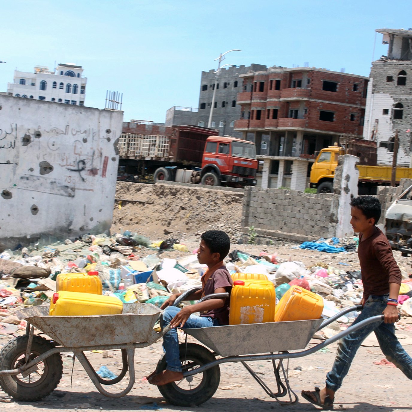 L'image montre deux enfants dans un environnement urbain en détérioration. L'un des enfants est assis dans une brouette, tandis que l'autre les pousse. Ils transportent des récipients jaunes, probablement pour l'eau, dans un paysage entouré de débris et de déchets. En arrière-plan, on peut voir des bâtiments en ruine et des camions. L'atmosphère semble difficile, reflétant une réalité de vie dans une zone touchée par des conflits ou des crises.