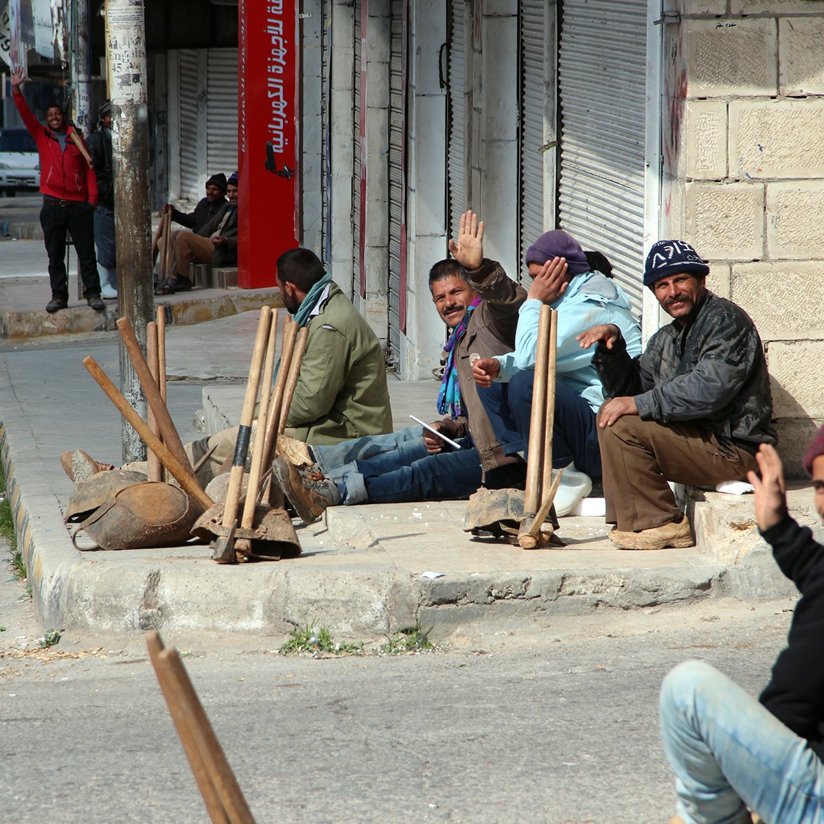 L'image montre un groupe d'hommes assis sur le bord de la route, apparemment en attente. Ils semblent discuter et certains saluent la caméra. L'environnement a l'air urbain, avec des bâtiments en arrière-plan et une rue. Des outils de travail sont visibles, suggérant qu'ils pourraient être des travailleurs. L'atmosphère semble décontractée malgré le cadre urbain.
