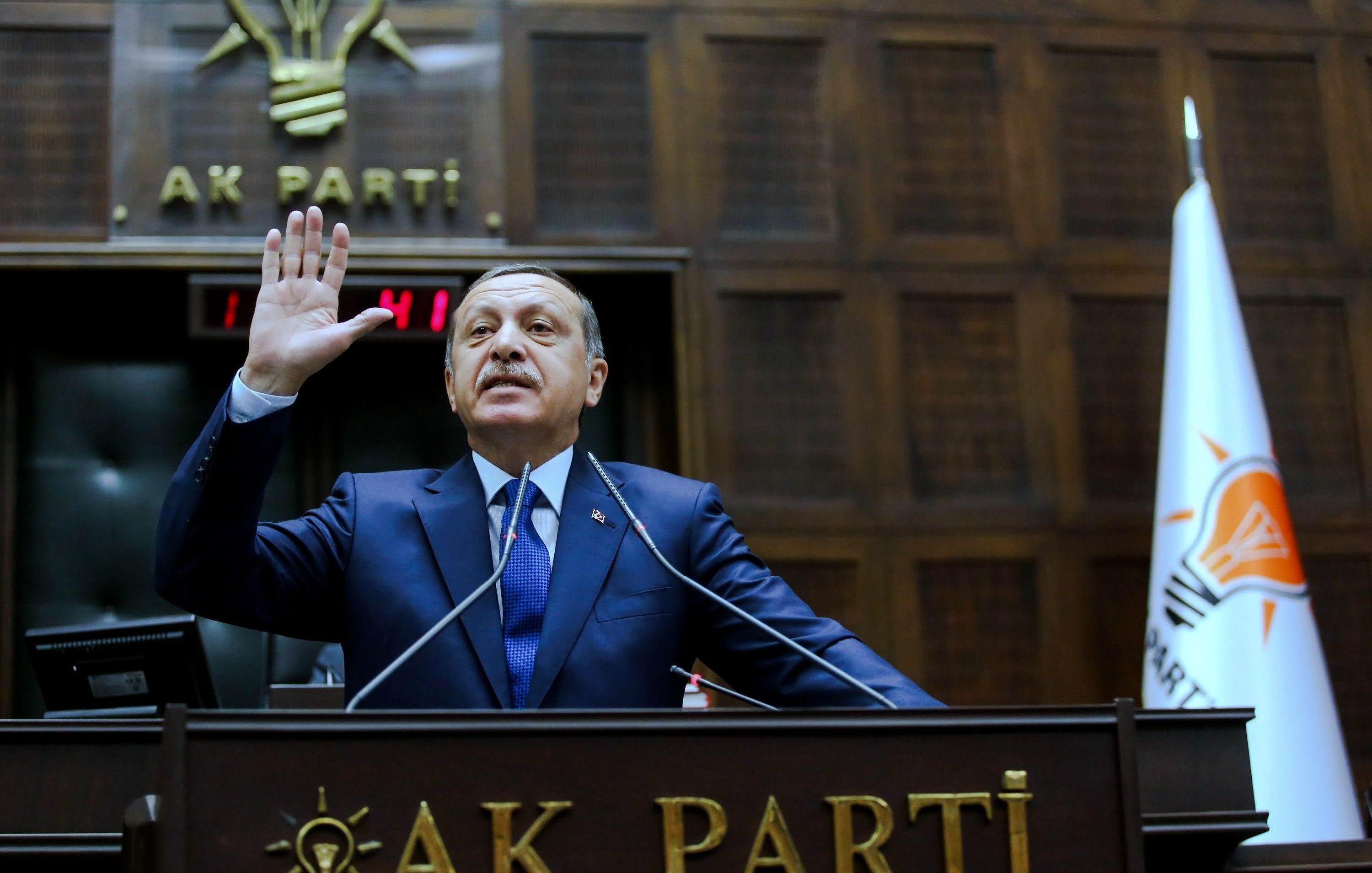 L'image montre un homme, probablement dans un contexte politique, levant la main en signe de serment ou de déclaration. Il est vêtu d'un costume sombre et d'une cravate. Derrière lui, on peut voir des éléments de décoration liés à un parti politique, avec un emblème et un drapeau. L'environnement semble être celui d'une assemblée ou d'un conseil, suggérant une atmosphère formelle et politique.