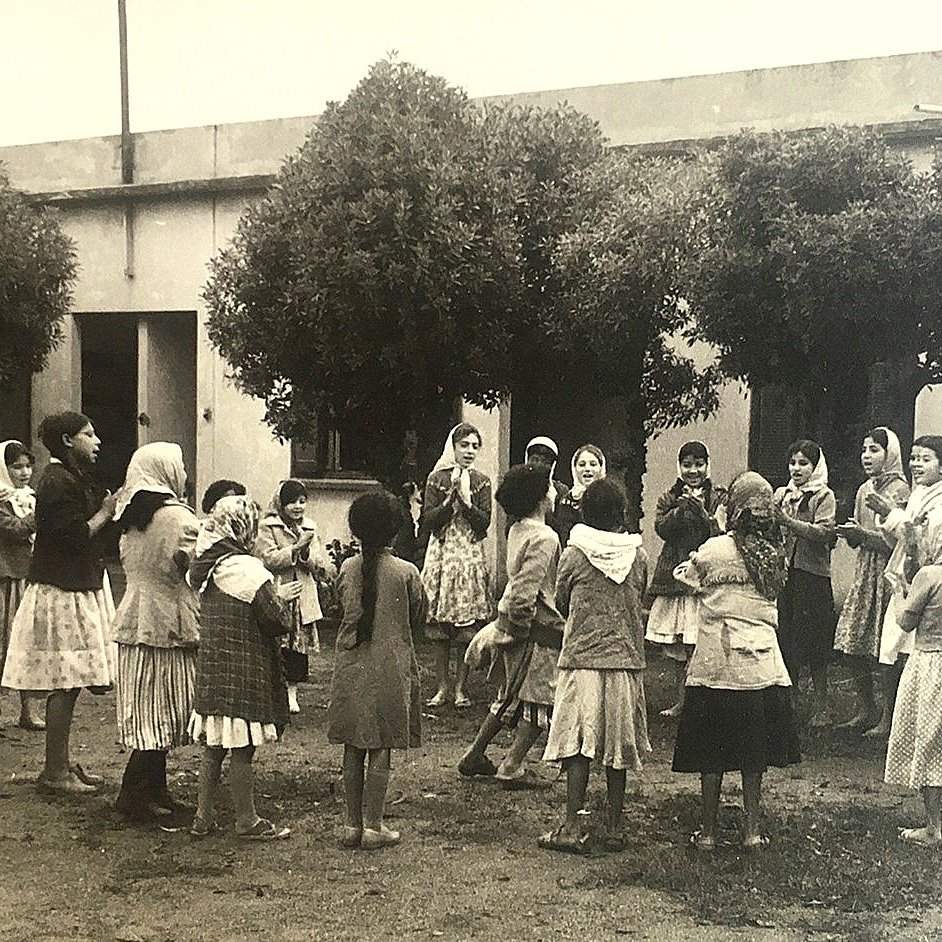 L'image montre un groupe d'enfants, principalement des filles, rassemblés dans une cour. Ils semblent jouer ensemble, entourés de grands arbres. Les filles portent des robes et certaines ont des foulards sur la tête. L'ambiance est joyeuse et festive, suggérant un moment de camaraderie et d'amusement. L'arrière-plan est constitué de murs d'un bâtiment, ce qui donne une atmosphère de communauté.