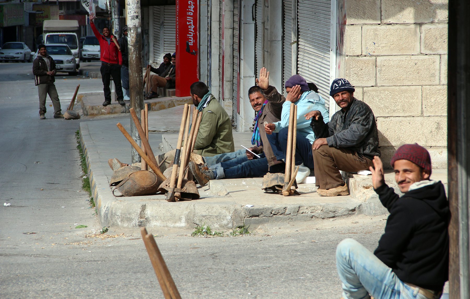 The image depicts a group of men sitting on a sidewalk in an urban setting. They appear to be relaxed, some waving at the camera, and are dressed in casual clothing. The street is relatively quiet, with a few vehicles in the background. There are also some construction tools and materials scattered around, suggesting they may be waiting for work or gathering. The environment looks somewhat desolate, with shuttered storefronts and a hint of a dusty road.