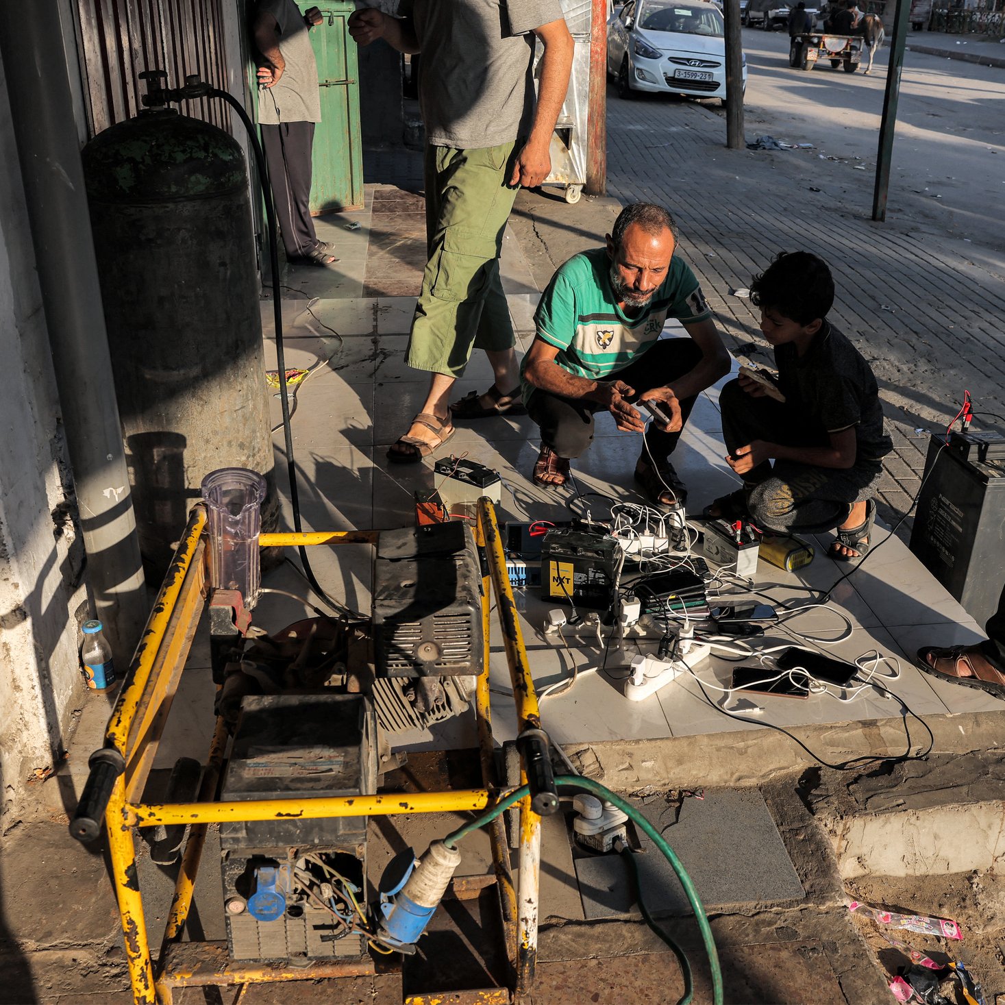 Nell'immagine si possono vedere alcune persone raccolte in un'area urbana. Tre uomini sono intenti a lavorare attorno a un dispositivo, probabilmente un generatore elettrico, che è posizionato su una base di cemento. Accanto al generatore ci sono vari cavi e dispositivi elettronici, suggerendo che stanno cercando di ricaricare batterie o alimentare apparecchi. Uno degli uomini è accovacciato mentre gli altri due stanno osservando o interagendo con i cavi. Sullo sfondo, si intravedono auto e strutture urbane tipiche. L'atmosfera sembra essere di lavoro e collaborazione nel contesto di un'area pubblica.