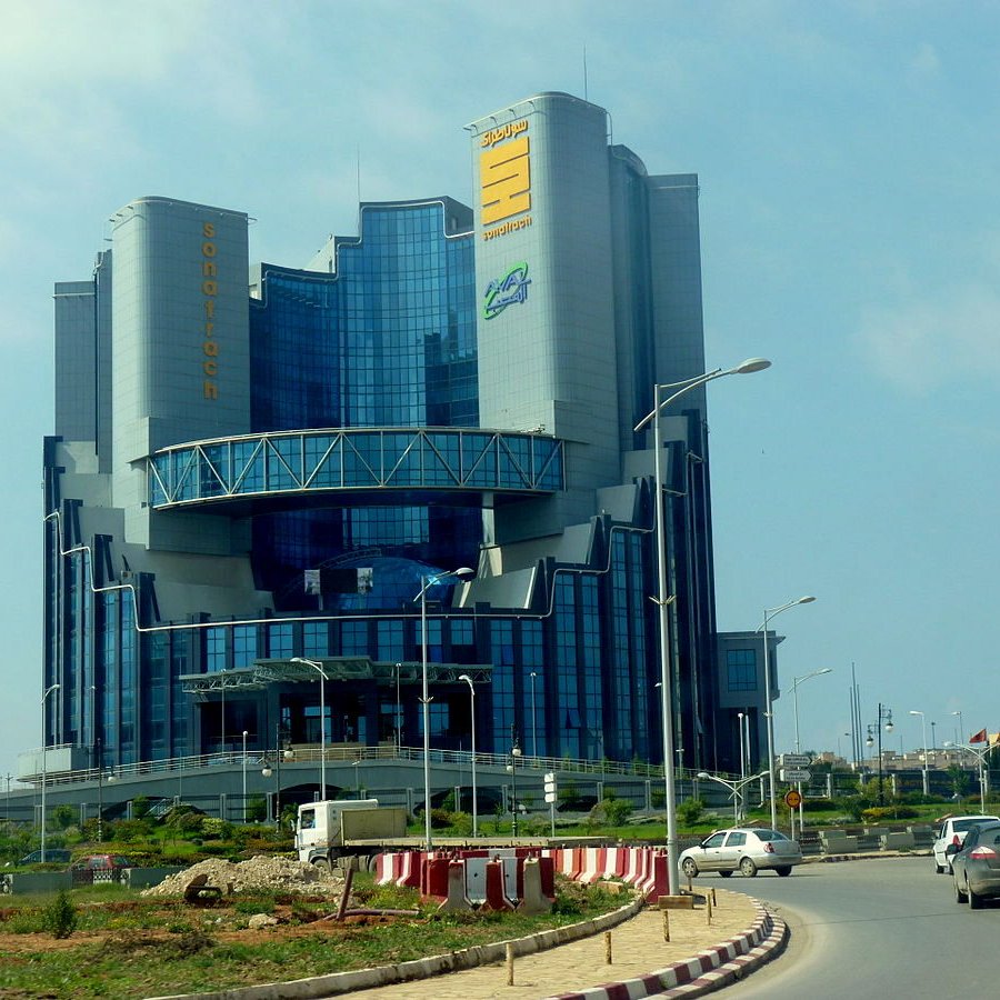L'image montre un bâtiment moderne et imposant, avec une architecture contemporaine. La structure se dresse sur plusieurs niveaux, avec de grandes fenêtres en verre qui reflètent la lumière. Au sommet, il y a un panneau avec des inscriptions en caractères distincts, suggérant que le bâtiment abrite une entreprise ou une organisation importante. Il est entouré d'une route et d'un espace vert, ce qui ajoute au cadre urbain. Le ciel est partiellement nuageux, ce qui donne un éclairage intéressant à l'ensemble de la scène.