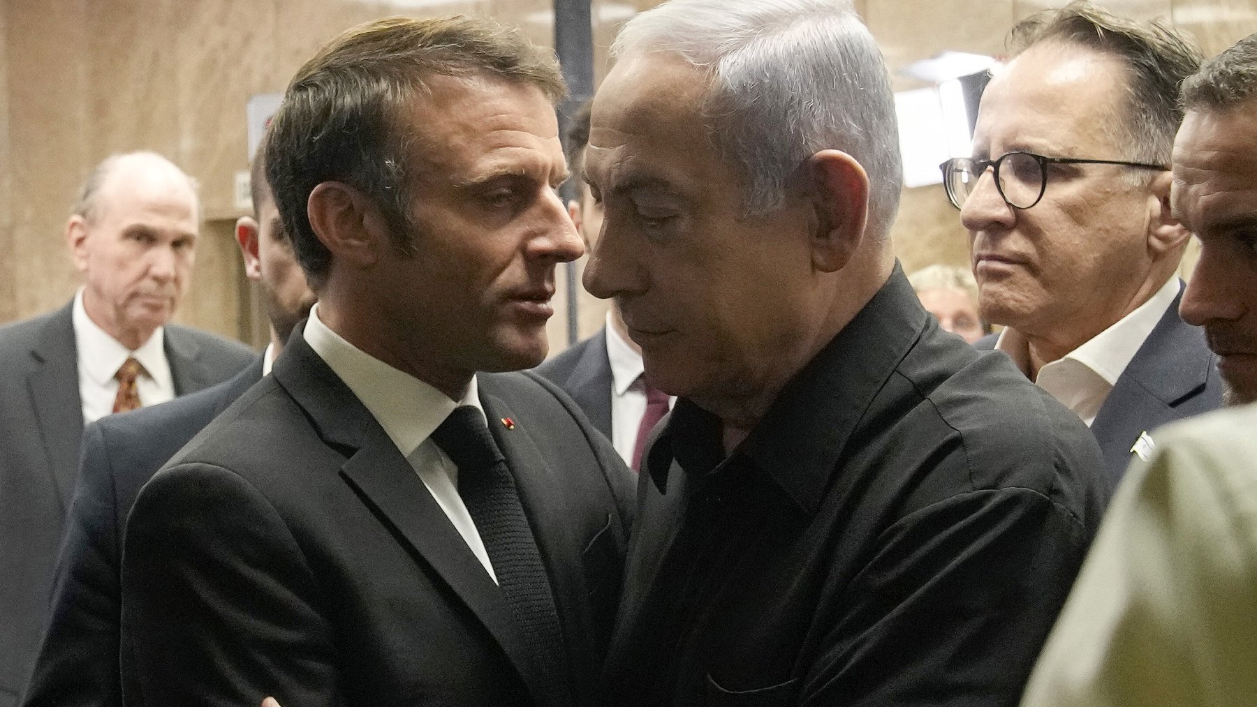 La imagen muestra a dos hombres en un entorno formal, aparentemente en un edificio gubernamental. Uno de ellos viste un traje oscuro y tiene una expresión seria mientras se acerca al otro, quien lleva una camisa negra. Ambos parecen en una conversación cercana, rodeados por varias personas que observan la interacción. La atmósfera sugiere un momento significativo, posiblemente de diálogo o entendimiento.