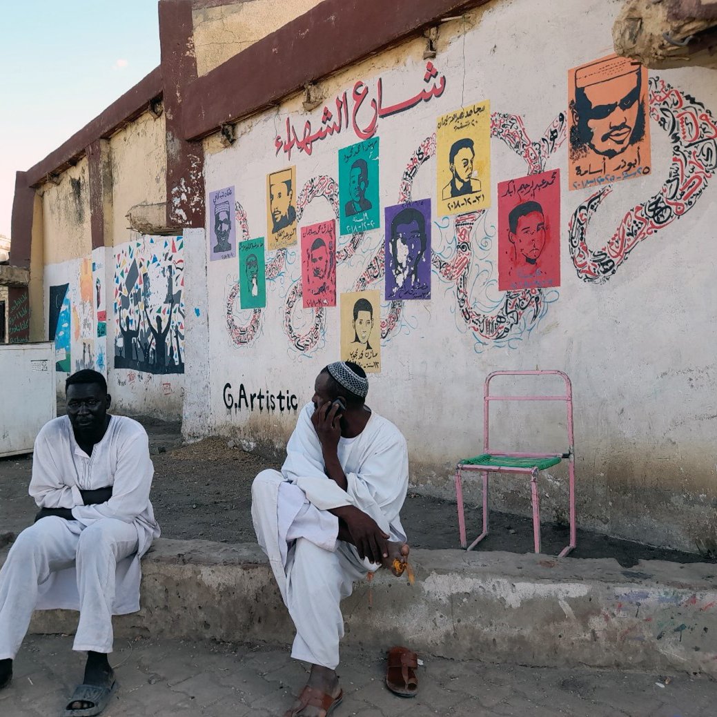 L'image montre deux hommes assis à l'extérieur, probablement dans une rue d'une ville. Ils portent des vêtements traditionnels, en particulier des robes longues. En arrière-plan, il y a un mur décoré de peintures murales colorées représentant des portraits. On peut également voir des motifs et des inscriptions en arabe. L'ambiance semble calme, avec des palmiers visibles en haut de l'image, suggérant une atmosphère chaleureuse.