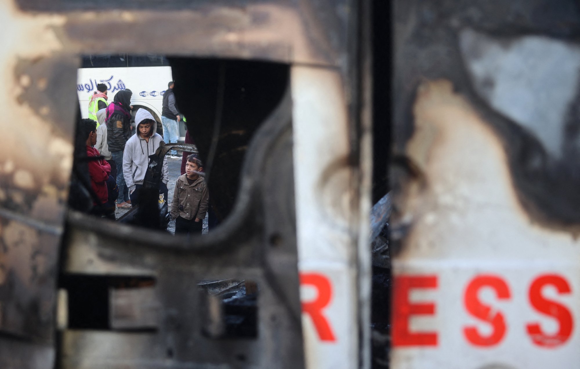 L'immagine mostra una scena cupa e difficile, probabilmente in un contesto di conflitto o crisi. Si vede un gruppo di persone che si trovano in un'area devastata, ricevendo assistenza o cercando di mettersi in salvo. Parte della scena è incorniciata da un oggetto danneggiato, che contribuisce a esprimere la tensione e la difficoltà della situazione. Le persone nel gruppo appaiono preoccupate e potrebbero essere in attesa di aiuto o di informazioni.