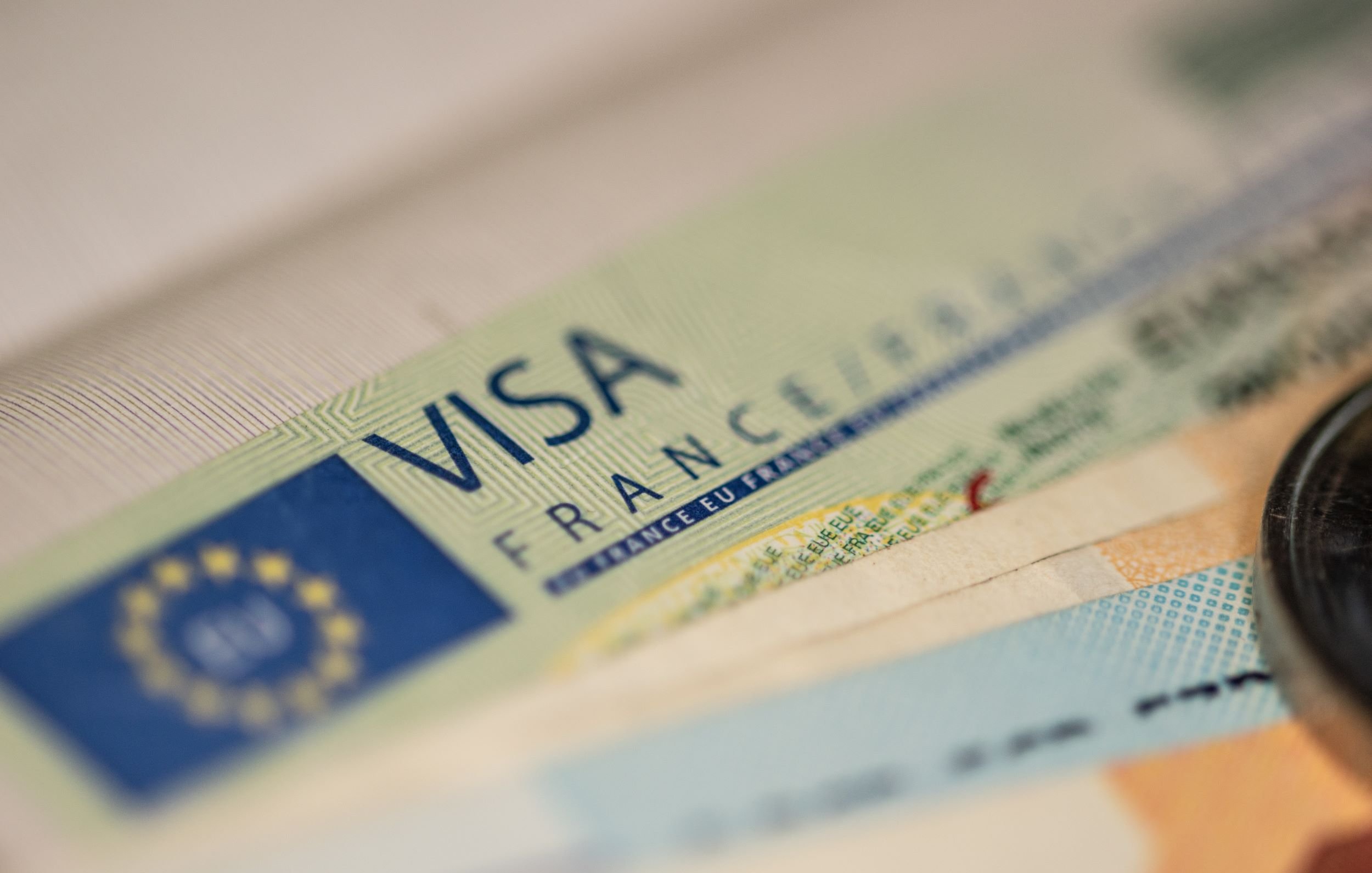 L'image montre un document de type visa, probablement un visa français. On peut voir le mot "VISA" en lettres majuscules, ainsi que des détails graphiques et textuels associés, comme le terme "FRANCE". En arrière-plan, on aperçoit des billets de banque, ce qui suggère un contexte lié à des voyages ou à des transactions. L'ensemble est présenté de manière à mettre l'accent sur le visa.