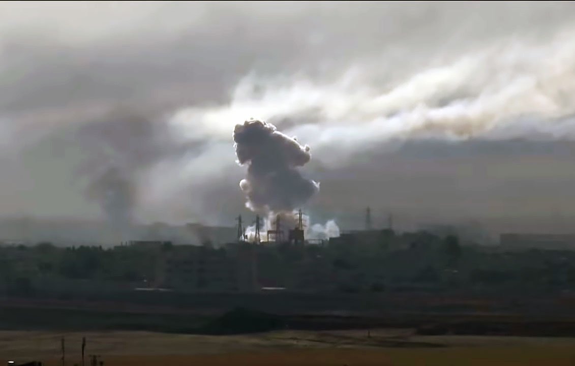L'image montre une scène dramatique avec des nuages de fumée épais s'élevant dans le ciel, probablement à la suite d'une explosion ou d'un bombardement. On peut voir des silhouettes d'infrastructures industrielles ou urbaines partiellement obscurcies par la fumée. L'atmosphère est sombre et chargée, suggérant des conditions de conflit ou de destruction. La lumière semble faible, ajoutant à l'impression de chaos et de détresse.