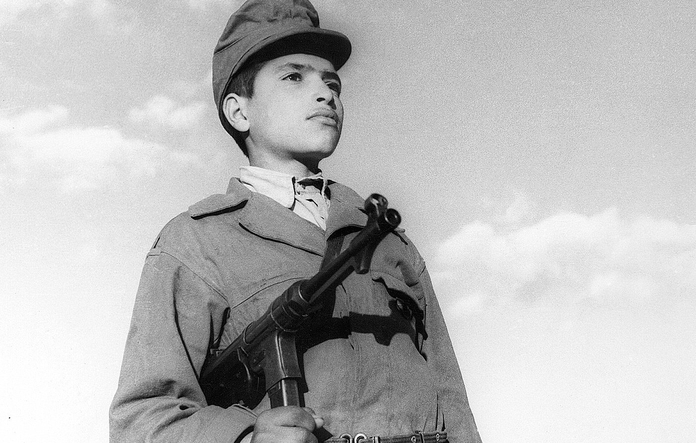 L'image montre un jeune garçon en uniforme militaire, tenant une arme à feu. Son regard est sérieux et déterminé, tandis qu'il se tient droit, face à l'objectif. Le ciel en arrière-plan est clair, avec quelques nuages. L'uniforme et l'arme peuvent indiquer qu'il fait partie d'un contexte militaire ou historique spécifique. L'ambiance de la photo évoque un sentiment de courage et de bravoure.