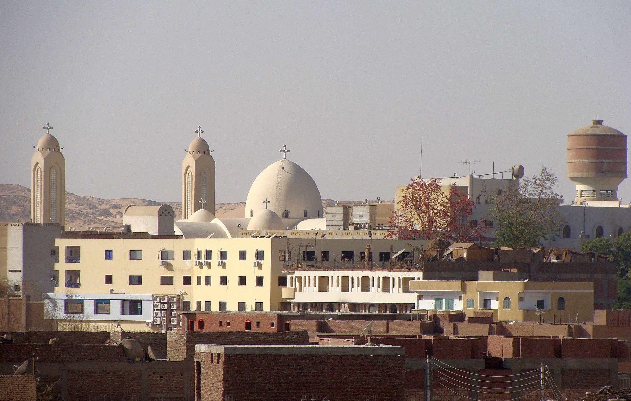 L'image montre un paysage urbain avec des bâtiments variés. On peut distinguer plusieurs églises avec des croix sur leurs dômes, caractéristiques de l'architecture religieuse. Des structures modernes cohabitent avec des bâtiments plus traditionnels, créant un contraste dans l'architecture. À l'arrière-plan, des collines ou des montagnes sont visibles, ajoutant une dimension naturelle au paysage. L'ensemble dégage une atmosphère de vie quotidienne dans une région marquée par son héritage culturel.