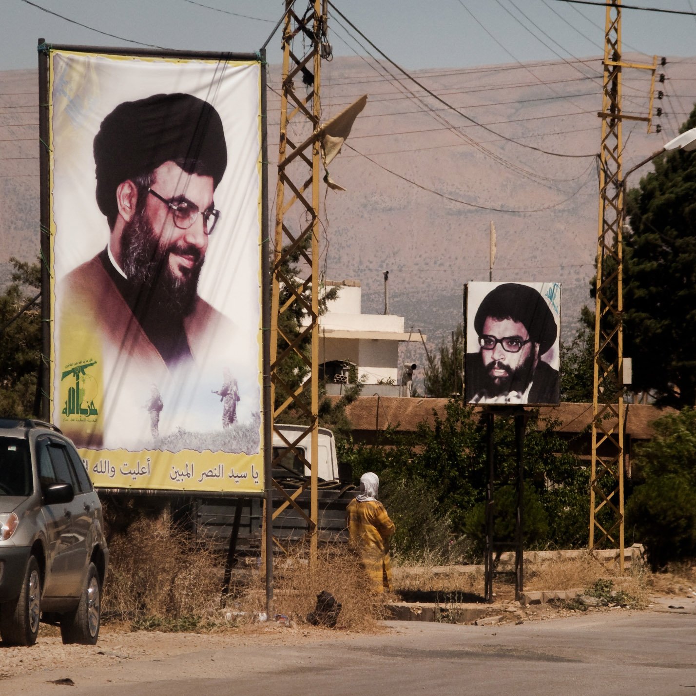 L'image montre une rue avec des panneaux d'affichage sur lesquels figurent des portraits d'un homme portant des lunettes et une barbe, associé à une ambiance rurale. On peut également voir des lignes électriques et quelques bâtiments en arrière-plan. À gauche, un panneau est en couleur et à droite, un autre est en noir et blanc. La scène semble être située dans une région montagneuse. C'est une illustration d'un espace public où des affiches politiques ou religieuses peuvent être observées.