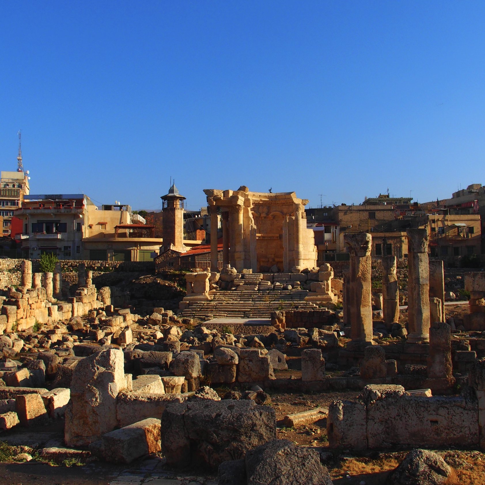 L'image montre un site archéologique avec des ruines anciennes, probablement d'un temple, entourées de colonnes et de blocs de pierre. En arrière-plan, on voit des bâtiments modernes et des structures urbaines s'élevant sous un ciel bleu. La lumière du soleil éclaire les ruines, créant un contraste entre l'ancien et le moderne. Ce lieu évoque une riche histoire et une importance culturelle.