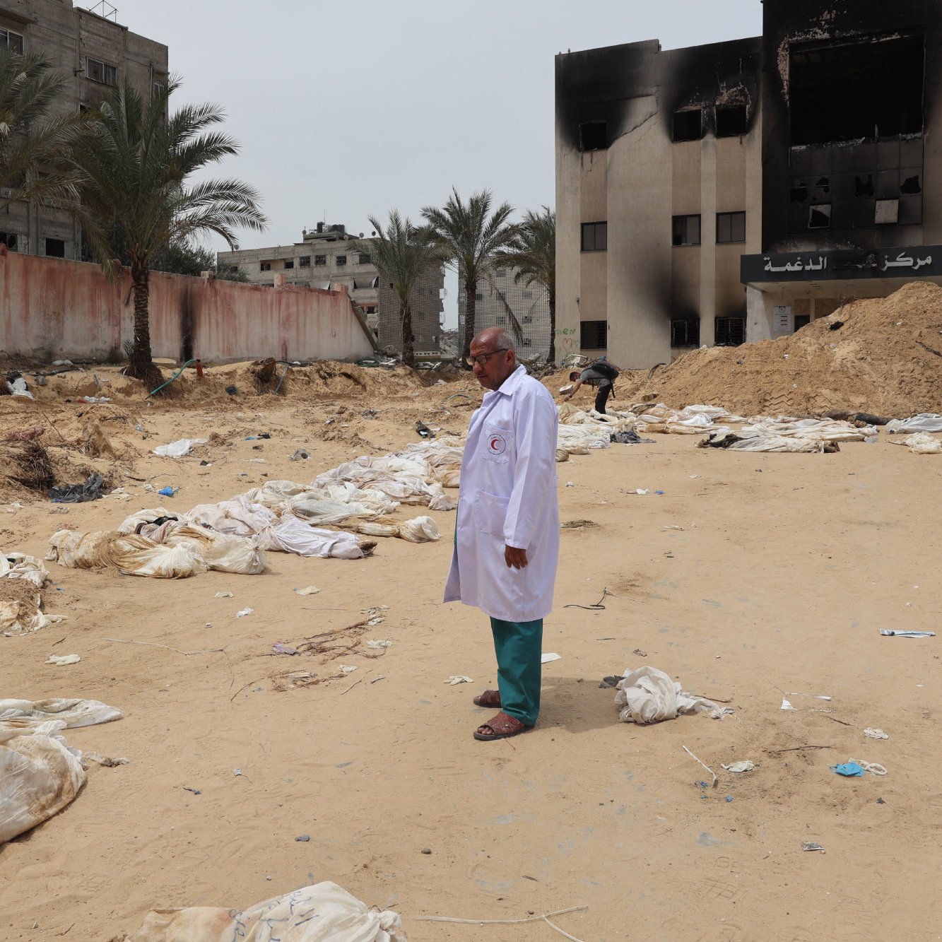 The image depicts a scene in a sandy area where several bodies, covered in white shrouds, are lying on the ground. In the foreground, a man wearing a white coat stands solemnly. In the background, there are damaged buildings, some of which appear to be burned. Palm trees line the area, and the overall atmosphere seems somber and grim, indicating a setting of conflict or disaster.