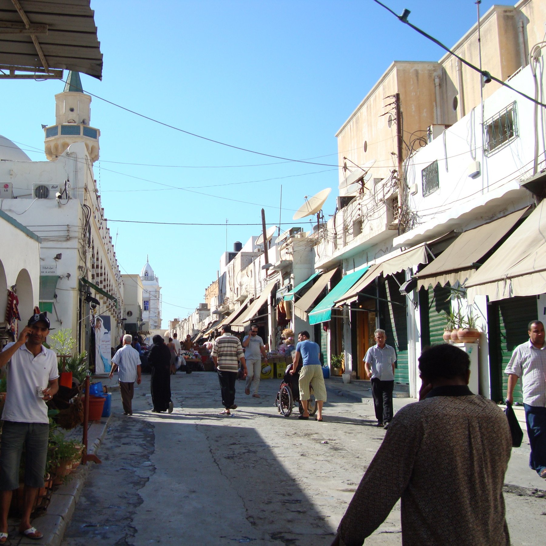 L'image montre une rue animée d'une ville typique, avec des bâtiments de style méditerranéen. On peut voir des personnes marchant le long de la rue, parmi lesquelles des hommes habillés en vêtements traditionnels. Des pots de fleurs et des végétaux décorent l'entrée de certains bâtiments, tandis que des ombrelles ou des auvents semblent offrir de l'ombre aux passants. Le ciel est dégagé et ensoleillé, créant une ambiance agréable.