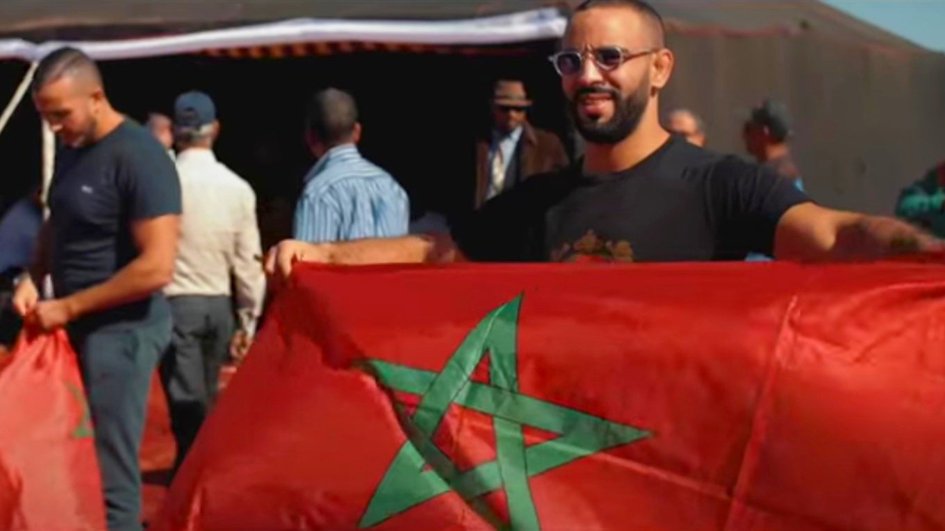 L'image montre un groupe de personnes, dont un homme tenant un drapeau marocain. Il sourit et affiche fièrement le drapeau rouge avec une étoile à cinq branches verte. En arrière-plan, on aperçoit d'autres personnes et des tentes, ce qui laisse supposer une rencontre ou un événement en plein air. L'atmosphère semble festive et conviviale, évoquant un rassemblement culturel ou patriotique.