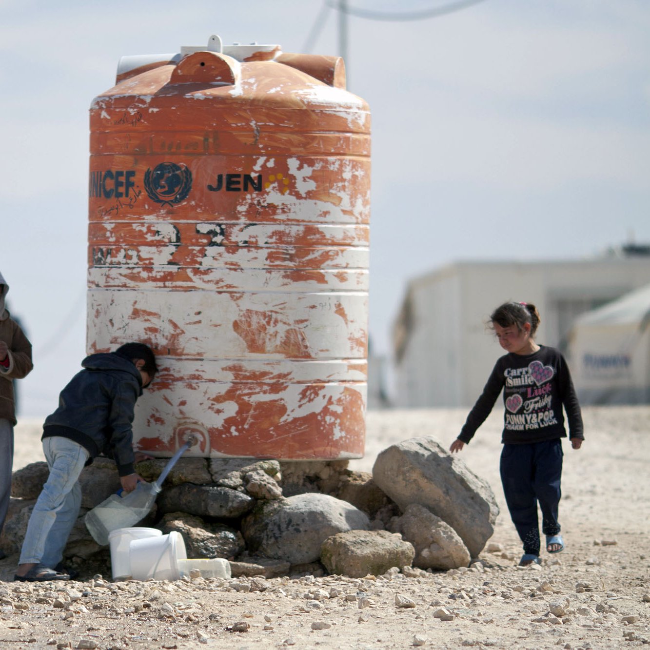 تظهر الصورة مجموعة من الأطفال بالقرب من خزان ماء كبير مُسجّل عليه علامة الأمم المتحدة. يقف اثنان من الأطفال بجانب الخزان، حيث يبدو أحدهم مشغولاً في فحص شيء ما، بينما يقوم الآخر بسكب الماء في وعاء. هناك طفل ثالث يلعب بجانبهم. الخلفية تبدو قاحلة مع بعض الصخور والأرض الرملية، مما يشير إلى بيئة جافة.