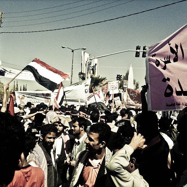 L'image montre une manifestation animée avec une grande foule de personnes. Des drapeaux flottent dans l'air, en particulier un drapeau qui semble être celui du Yémen. On peut voir des hommes et des femmes rassemblés avec des expressions déterminées. Des pancartes sont tenues par des manifestants, certaines comportant des inscriptions en arabe. L'atmosphère est dynamique, reflétant le désir de changement ou de revendication au sein de la foule. Les bâtiments environnants et les lignes électriques en arrière-plan ajoutent un contexte urbain à la scène.
