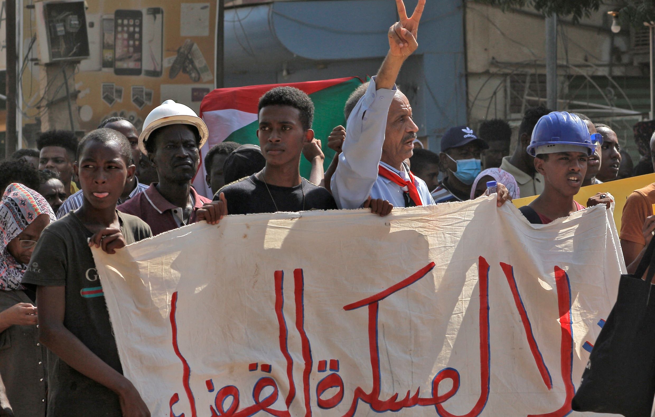 L'image montre une scène de manifestation, probablement en rapport avec des revendications politiques. On y voit des personnes tenant une grande bannière. Un homme au premier plan fait un signe de paix avec ses doigts. Les manifestants semblent déterminés et engagés, et des drapeaux sont visibles en arrière-plan, indiquant un contexte national. L'atmosphère est chargée d'émotion, illustrant un moment de solidarité.