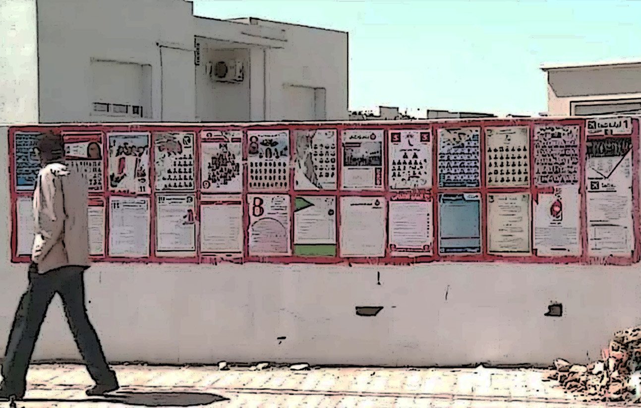 L'image montre un mur recouvert de plusieurs affiches et panneaux d'information. Ces affiches semblent contenir des annonces, des avis ou des informations diverses, affichées de manière colorée. Un homme marche devant ce mur, ce qui donne une idée de l'échelle des affiches et de l'environnement. Le paysage est lumineux, suggérant une journée ensoleillée.