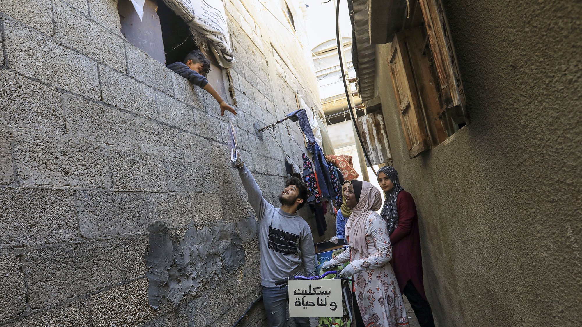 الصورة تُظهر مشهداً في زقاق ضيق حيث يبدو شاب يحمل شيئاً بيده وهو يمده نحو طفل يطل من نافذة في الجدار. في الجهة الأخرى، توجد امرأة ترتدي حجاباً متواجدة بجانب امرأة أخرى، وكلاهما تراقبان الموقف. يمكن رؤية أشياء معلقة في الجوار، مثل الملابس، وهناك دراجة ملونة موضوعة بجانب الجدار. الأجواء تعكس الحياة اليومية في منطقة مكتظة.