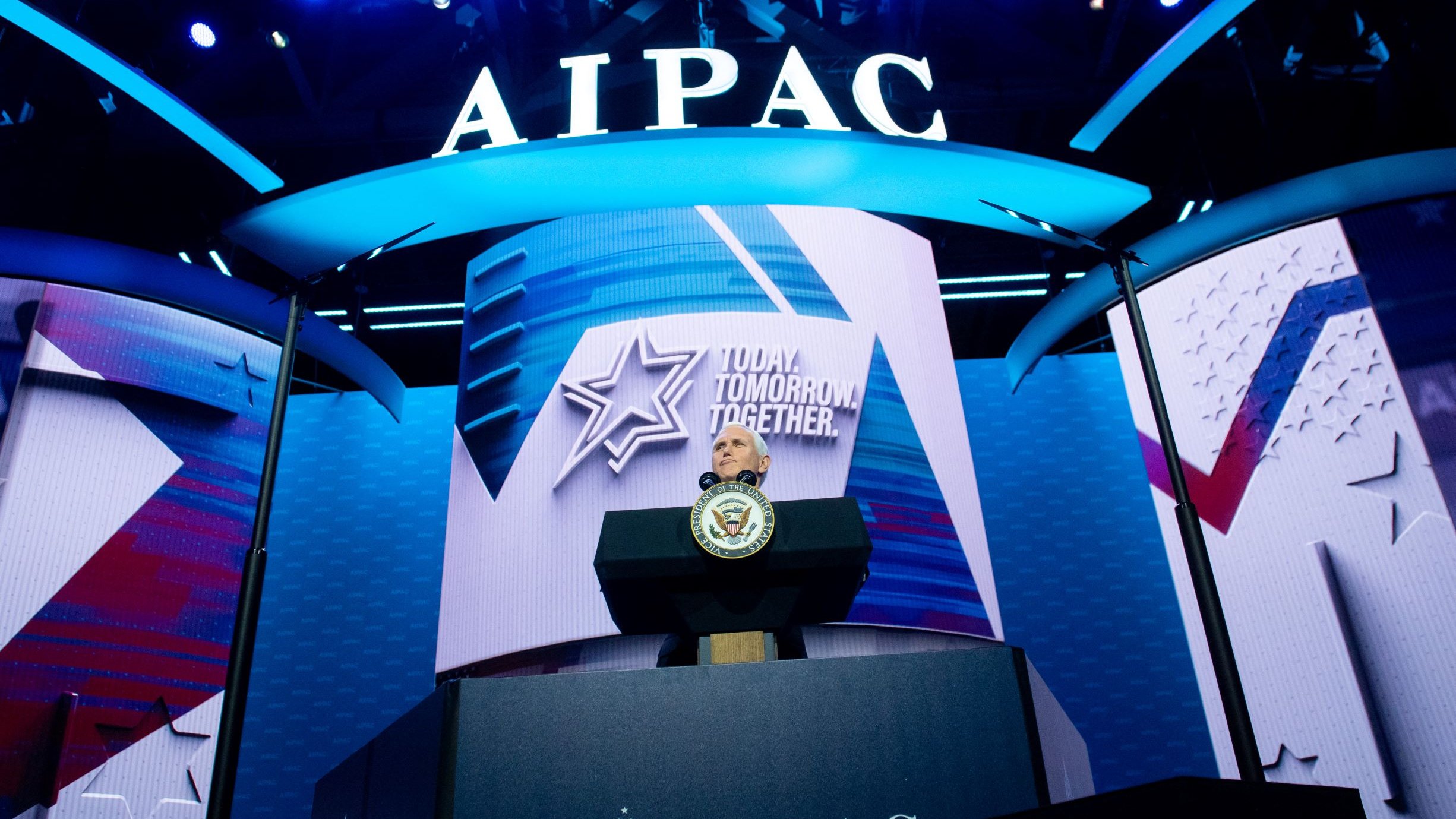La imagen muestra un escenario iluminado con luces brillantes en un evento, con un gran fondo donde se puede ver el logo de AIPAC (Comité de Relaciones Públicas América-Israel). Hay un podio en el centro, probablemente destinado a un orador, con un sello oficial, indicando que se trata de un evento importante. La decoración incluye elementos gráficos como estrellas y símbolos que refuerzan un mensaje de unidad y futuro, destacado en el fondo.