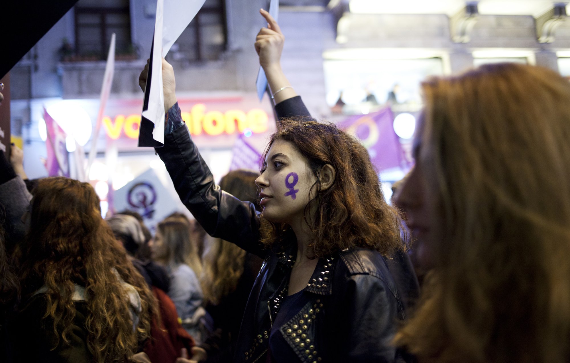 L'image montre un groupe de personnes participant à une manifestation. Au premier plan, une jeune femme avec des cheveux bouclés et un maquillage marqué, porte une veste en cuir et tient un morceau de papier ou une affiche. Son visage est décoré d'un symbole féminin en violet, qui se retrouve également sur d'autres bannières en arrière-plan. L'atmosphère semble être celle d'un rassemblement pour les droits des femmes, soulignant la solidarité et l'engagement des participants.