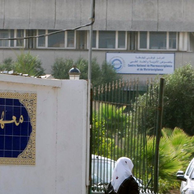 The image depicts a building with signage indicating it is the "Institut Pasteur" in Algeria. In the foreground, there is a decorative wall with Arabic text, likely the name of the institution, along with some greenery. You can also see several people walking along a path near the entrance. The architectural style of the building appears modern and utilitarian.