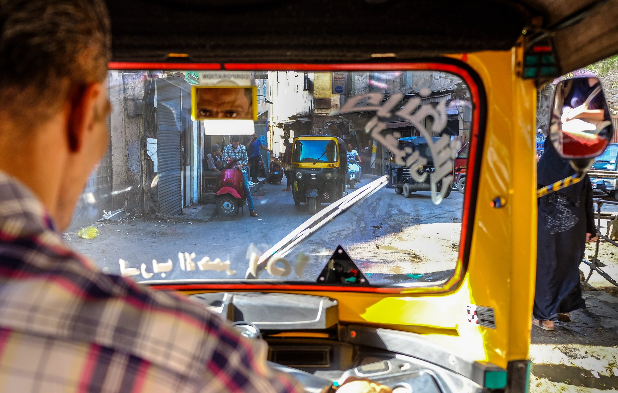 L'image montre la vue depuis l'intérieur d'un tuk-tuk, avec un conducteur visible en premier plan. On aperçoit des rues étroites et animées, avec d'autres véhicules, y compris un autre tuk-tuk. Des bâtiments anciens bordent la rue, et il y a des détails comme des affiches et des éléments de la vie urbaine qui ajoutent du caractère à l'environnement. La lumière naturelle éclaire la scène, créant une atmosphère vivante et dynamique.