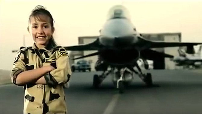 L'image montre une jeune fille souriante vêtue d'une tenue militaire camouflage. Elle se tient debout, les bras croisés, devant un avion de chasse. L'arrière-plan est flou, mettant l'accent sur l'avion, qui semble être un F-16. La scène dégage une ambiance de fierté et de bravoure, avec un ciel clair à l'horizon.