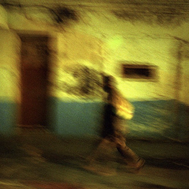 L'image présente une scène nocturne floue, où une silhouette se déplace le long d'un mur aux teintes décolorées, combinant des nuances de vert et de bleu. La texture du mur semble usée et vieillie, suggérant un environnement urbain. La personne, portant un sac, marche rapidement, créant un effet de mouvement qui renforce l'impression de dynamisme et d'évasion dans cette ambiance mystérieuse.
