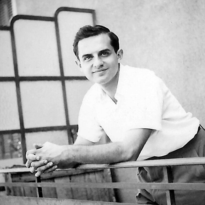 الصورة تظهر شابًا واقفًا على شرفة، يبدو مبتسمًا ومسترخيًا. يرتدي قميصًا أبيض وبنطالًا طويلًا. الخلفية تتضمن حائطًا وبعض التفاصيل المعمارية مثل النوافذ والحواجز. تعكس الصورة أجواءً من السعادة والبساطة.