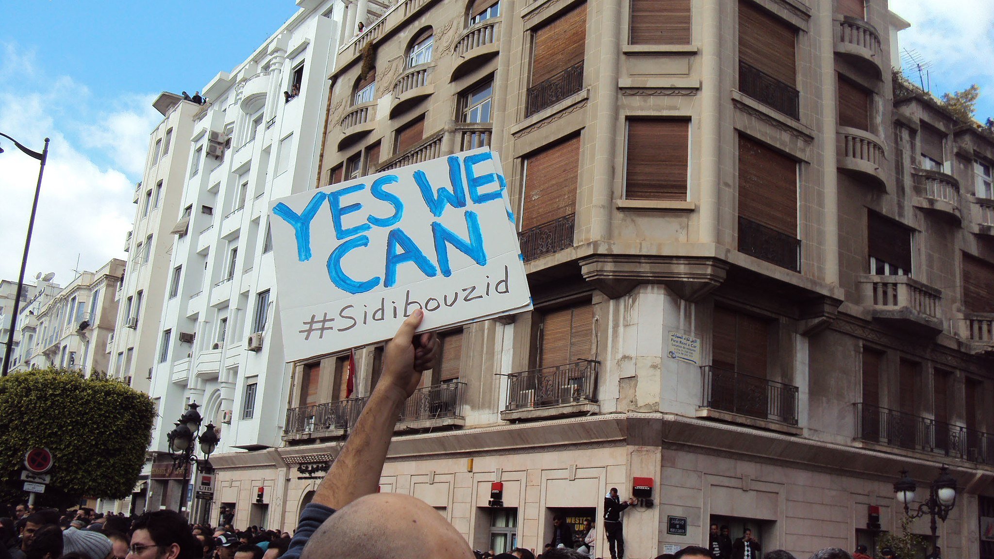 در این تصویر، گروهی از مردم در یک تجمع یا تظاهرات شرکت کرده‌اند. شخصی در جلوی تصویر، تابلویی با نوشته «YES WE CAN» و هشتگ «#Sidibouzid» را در دست دارد. ساختمان‌هایی با معماری مشخص در پس‌زمینه دیده می‌شود و جمعیت به‌نظر معترض یا فعال است. فضای تصویر حالت اجتماعی و سیاسی به خود گرفته و نشان‌دهندهٔ یک حرکت جمعی است.