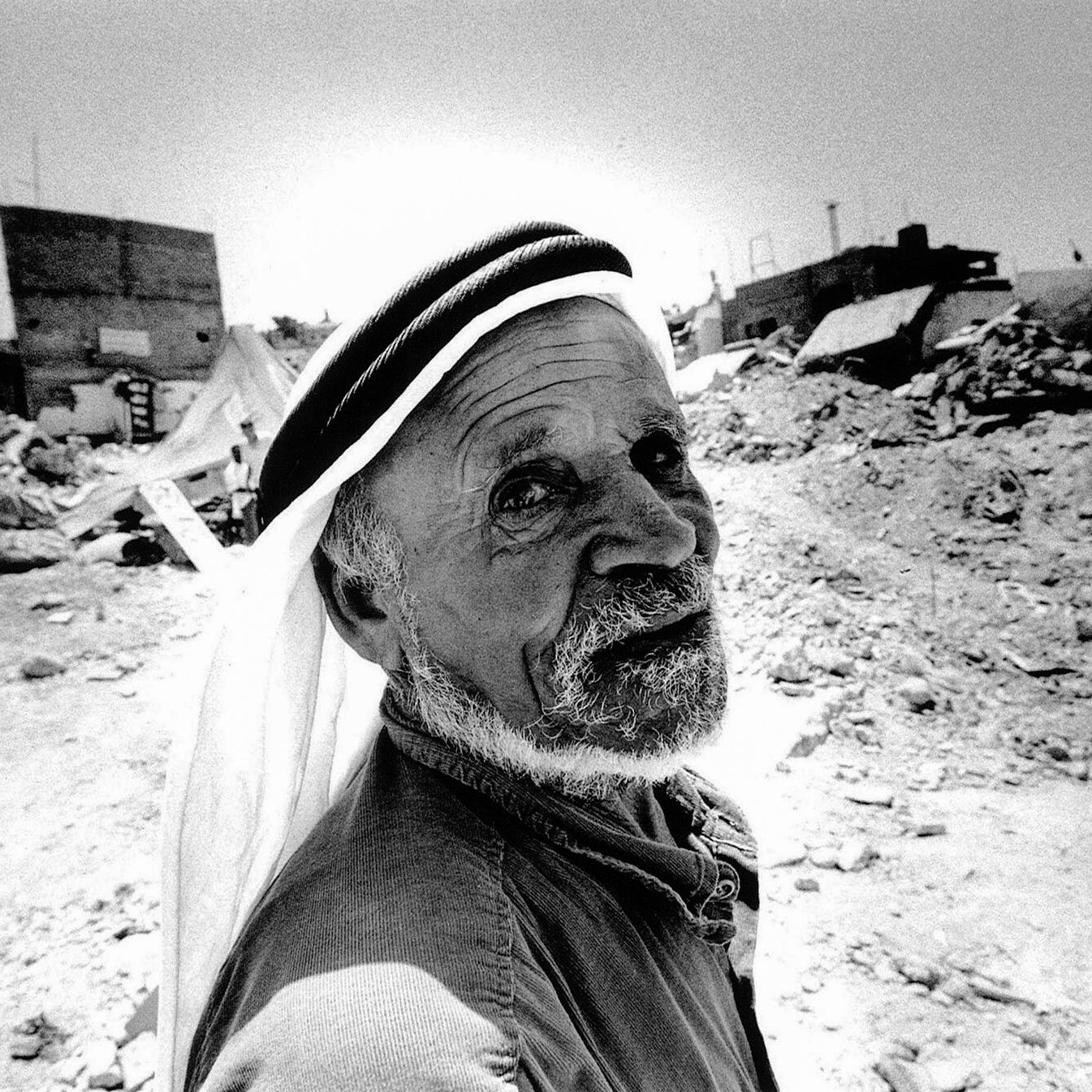 L'image montre un homme âgé au visage marqué, qui regarde directement vers l'objectif. Il porte un keffieh, un couvre-chef traditionnel, et a une expression qui dégage une certaine sagesse ou tristesse. En arrière-plan, on peut apercevoir des ruines, des débris et des bâtiments partiellement détruits, suggérant un environnement difficile ou une situation de conflit. La scène est capturée en noir et blanc, ce qui accentue les textures et les contrastes des éléments environnants.