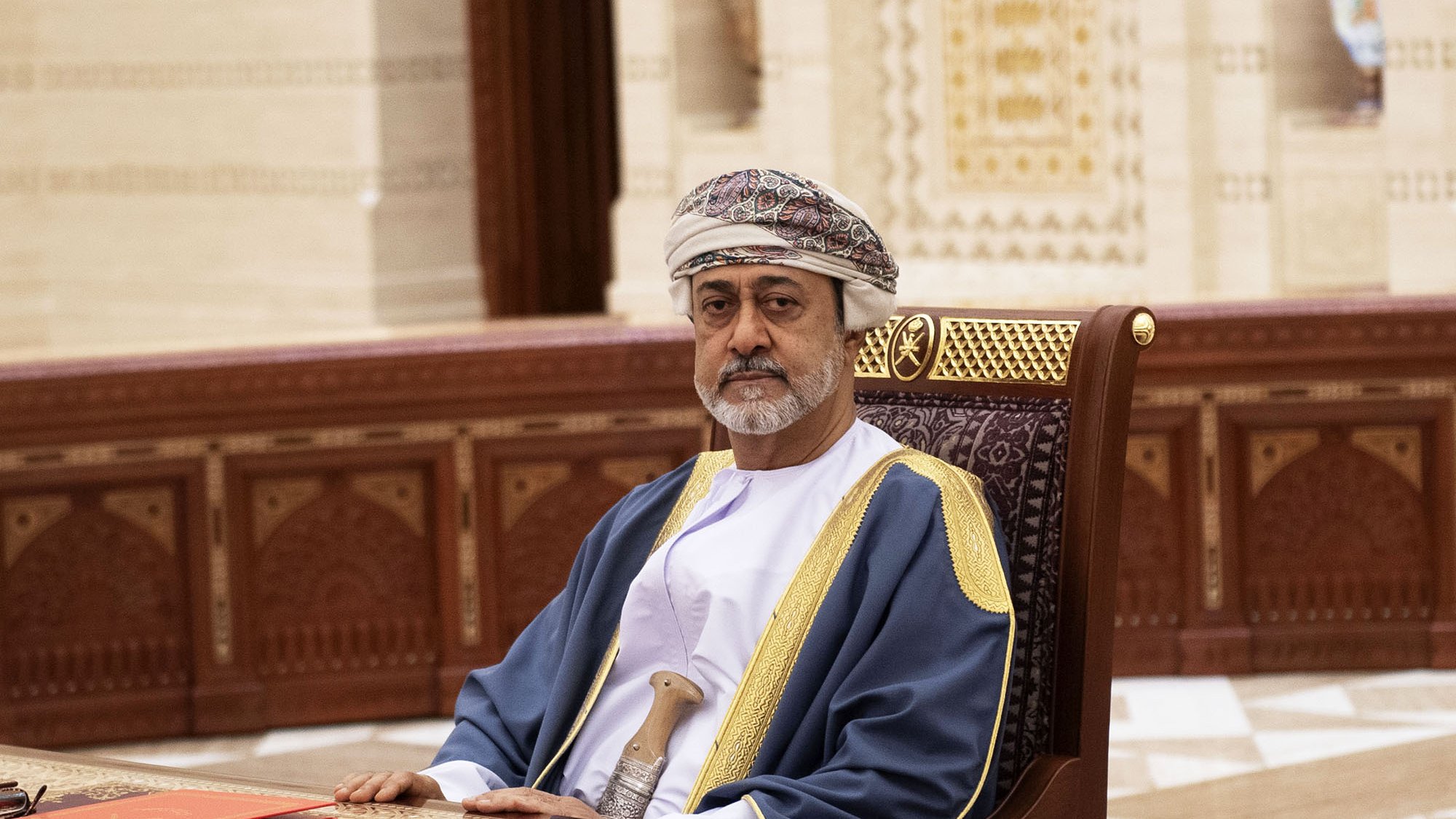 L'image montre un homme assis à un bureau dans un environnement intérieur richement décoré. Il porte un costume traditionnel, comprenant une robe longue et un turban orné. Le décor autour de lui présente des motifs architecturaux détaillés, typiques d'une ambiance élégante. Il semble concentré et sérieux, et le mobilier en bois ajoute à l'atmosphère formelle de la scène.