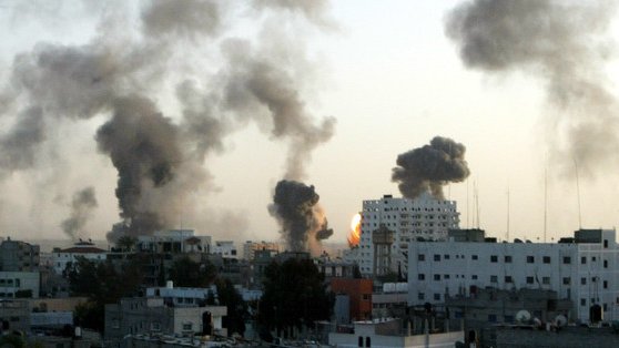 L'image montre une vue d'une ville en ruines, avec plusieurs colonnes de fumée s'élevant dans le ciel au crépuscule. Les bâtiments sont partiellement détruits, et l'atmosphère semble tendue et chaotique. La lumière du coucher de soleil se mêle aux nuages de fumée, créant un contraste marqué entre la lumière et l'obscurité. Cela évoque une scène d'urgence ou de conflit.