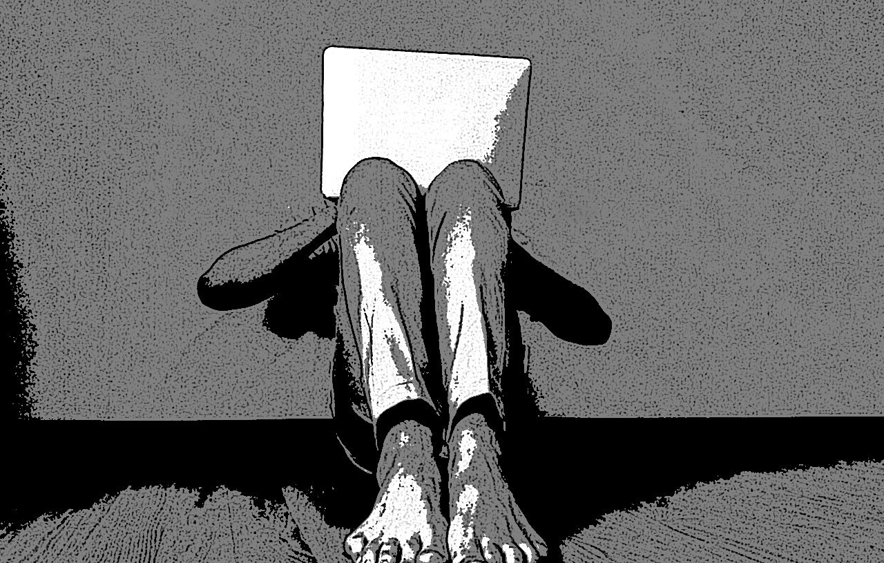 L'image montre une personne assise contre un mur, les jambes repliées et les pieds nus visibles. Elle tient un ordinateur portable ouvert devant elle, cachant probablement son visage. Le contraste noir et blanc donne un aspect artistique et minimaliste à la scène. Le fond est simple et met en avant la posture de la personne, ajoutant une impression de solitude ou d'introspection.